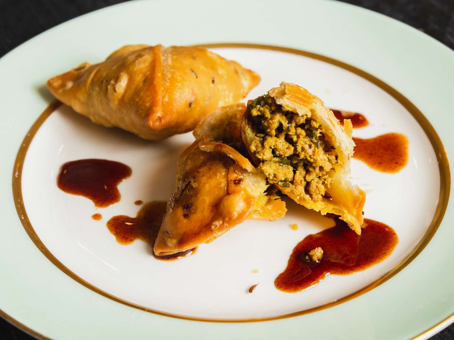 Two kheema samosas on a plate, one broken open to show its interior.