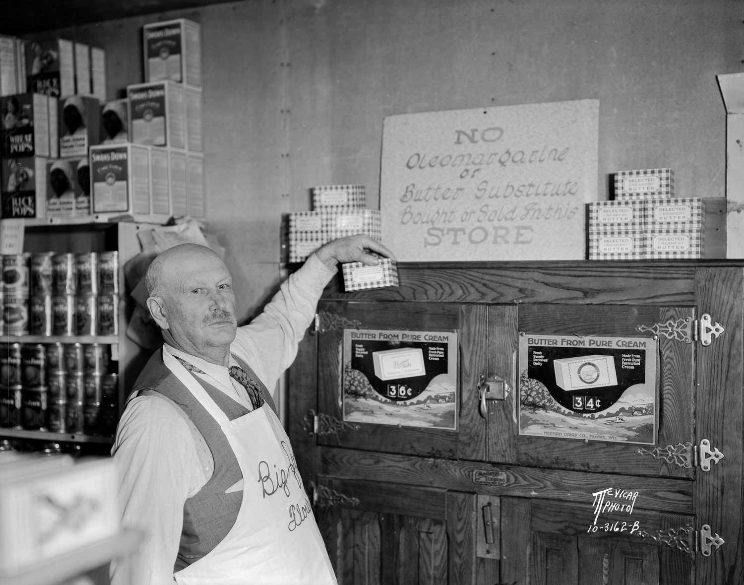 A man wearing an apron standing near a cabinet and pointing to a sign prohibiting the sale of colored margarine