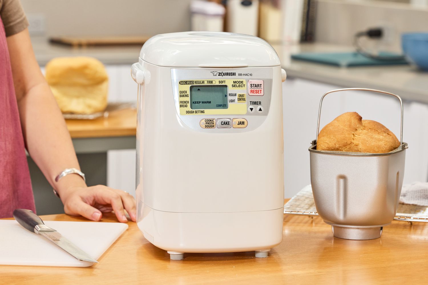 The Zojirushi Home Bakery Mini Breadmaker on a wooden counter next to a basket filled with bread