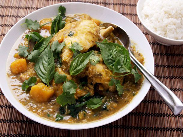 An overhead view of a white bowl of Thai green chicken curry with eggplant and squash, garnished with fresh basil next to a bowl of rice.