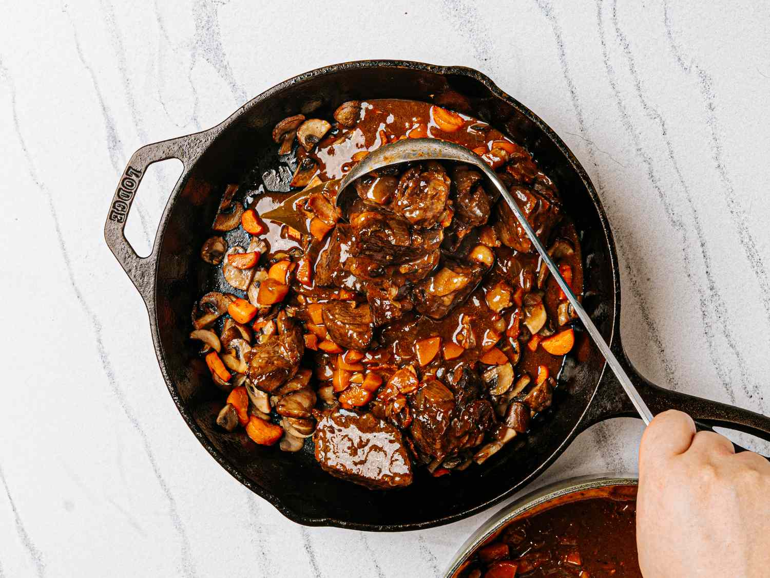 A hand stirring beef bourguignon in a skillet with a ladle the dish includes beef chunks and vegetables in rich gravy