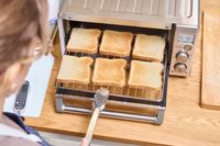 A person using tongs to slide out the rack of the Breville the Compact Smart Oven containing 6 slices of toast