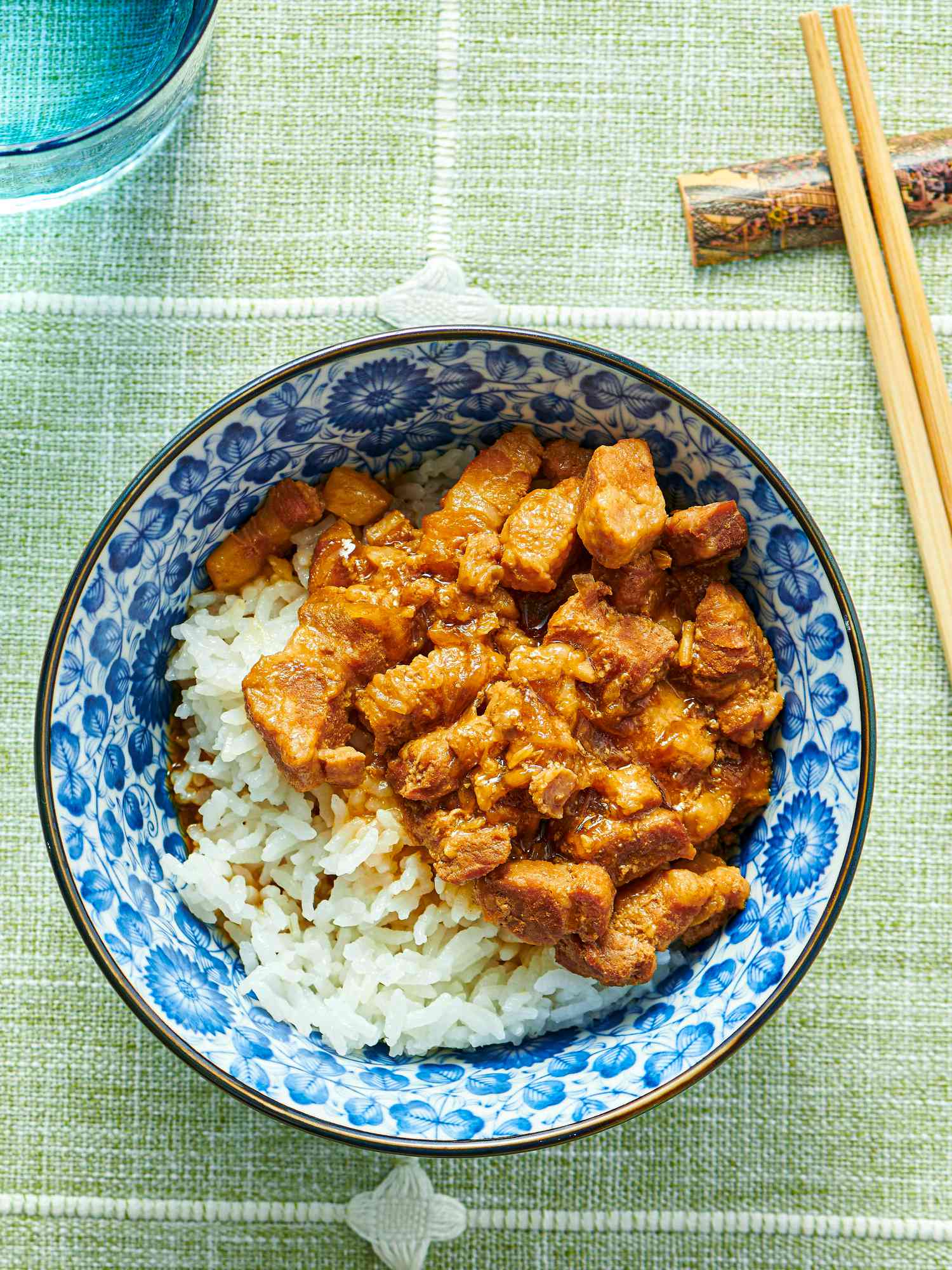 pressure cooked minced pork on white rice, in a blue bowl. Chopsticks and blue glass of water, on green tablecloth