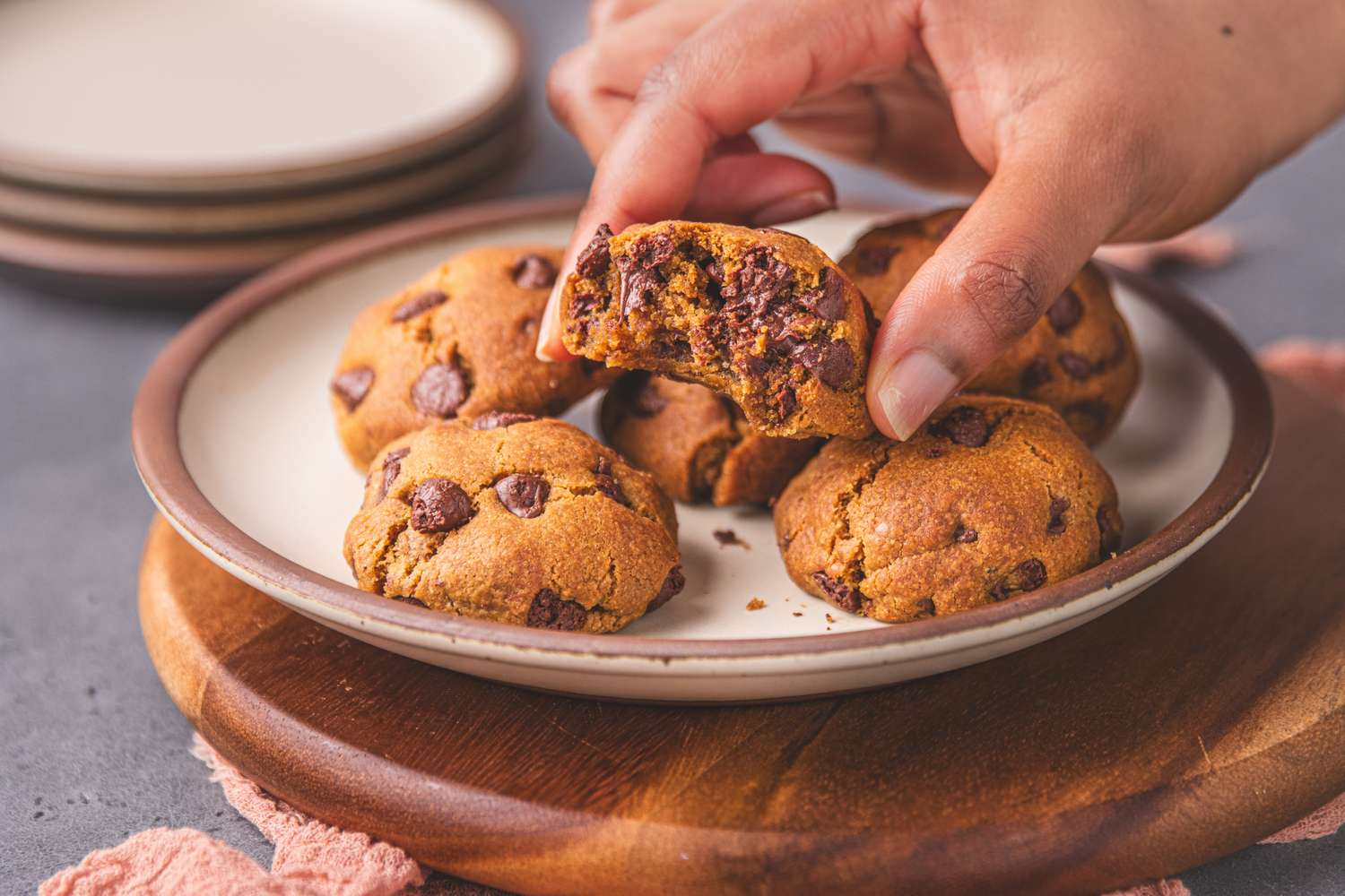 A plate of chocolate chip cookies with a hand holding one cookie to show the inside texture