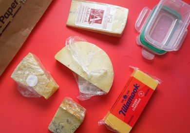 a variety of cheese on a red backdrop with parchment paper and a food container