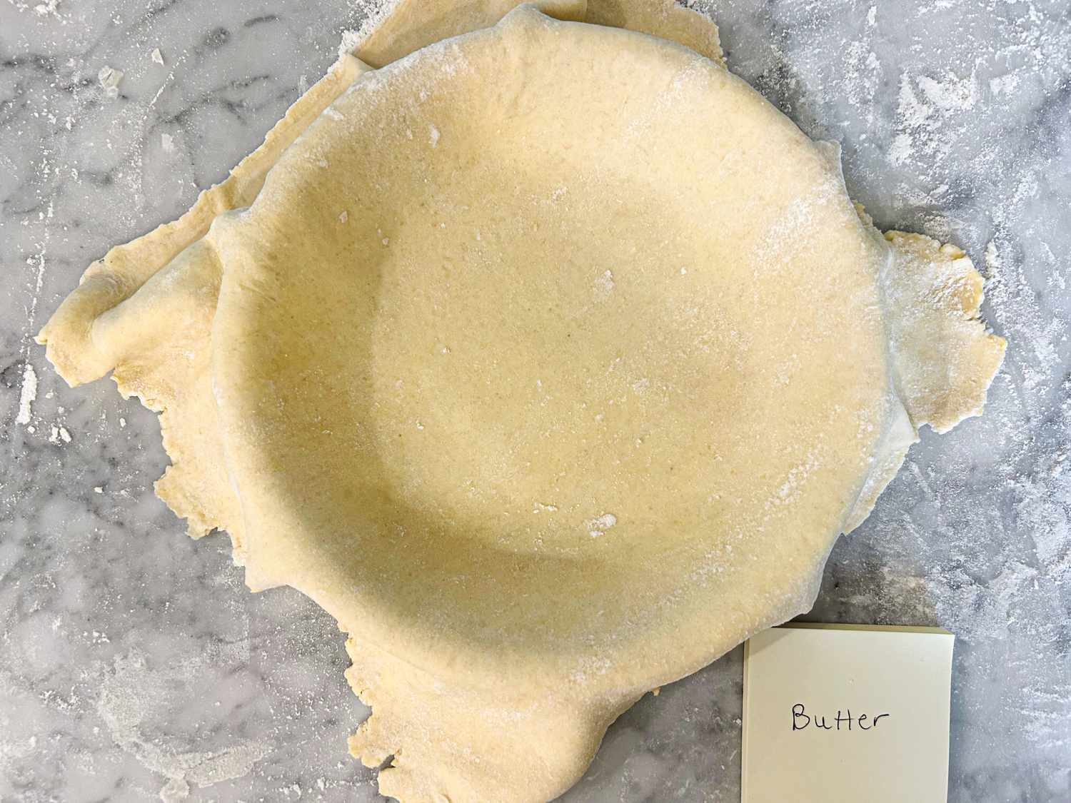 A pie crust in a dish on a floured surface with a note labeled butter nearby