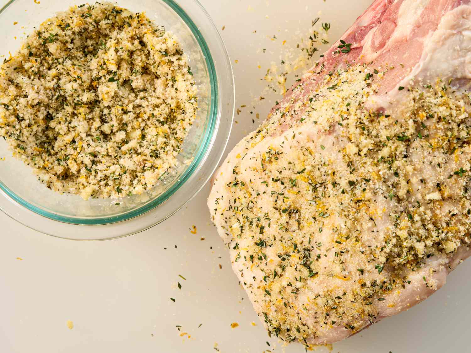 A fresh ham coated with herb seasoning next to a bowl of the mixture
