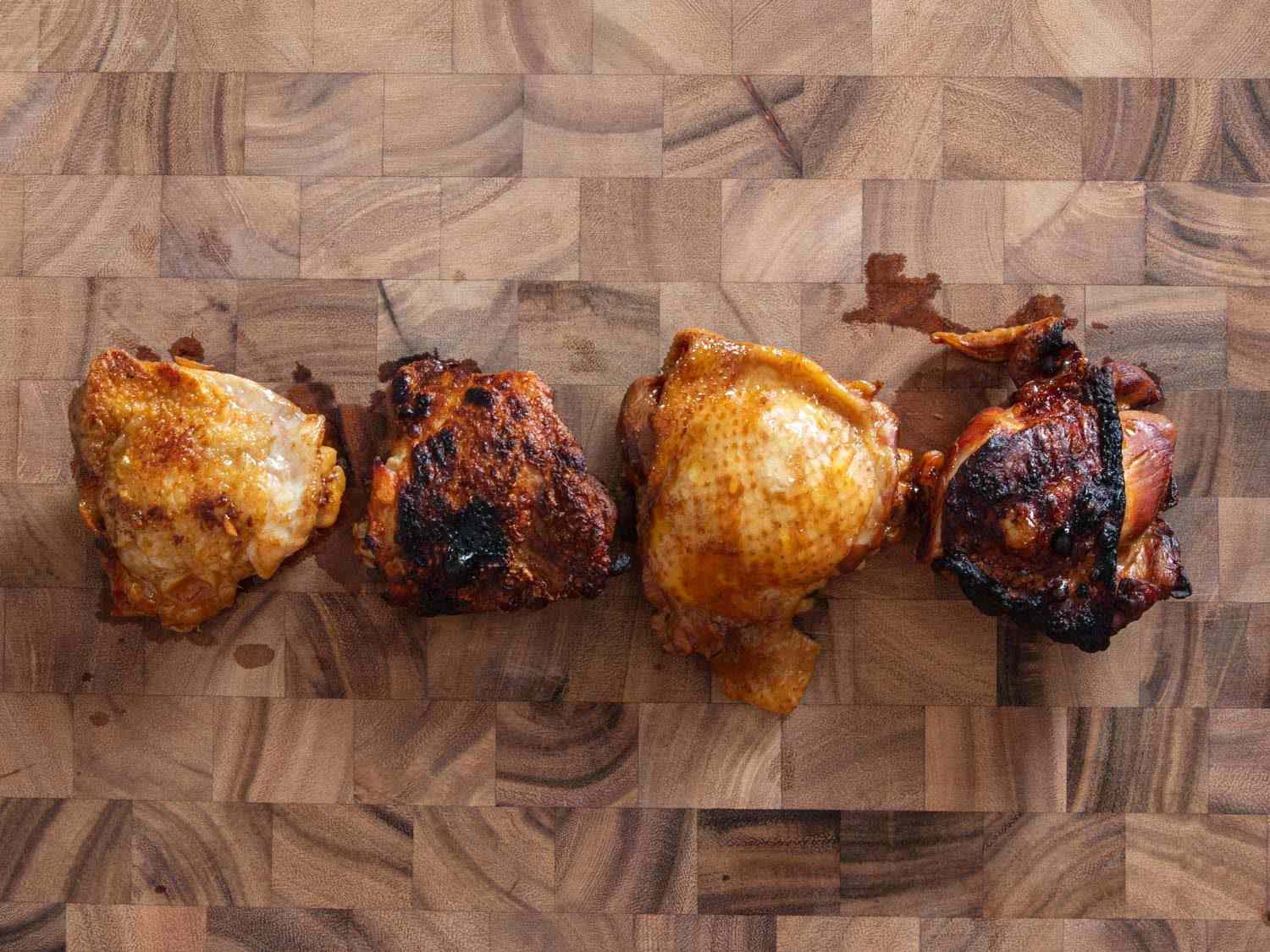 Side-by-side comparison of chicken thighs adobo on a cutting board. From left to right: Browned braised chicken thigh; browned braised and broiled chicken thigh; unbrowned braised chicken thigh; unbrowned braised and broiled chicken thigh.