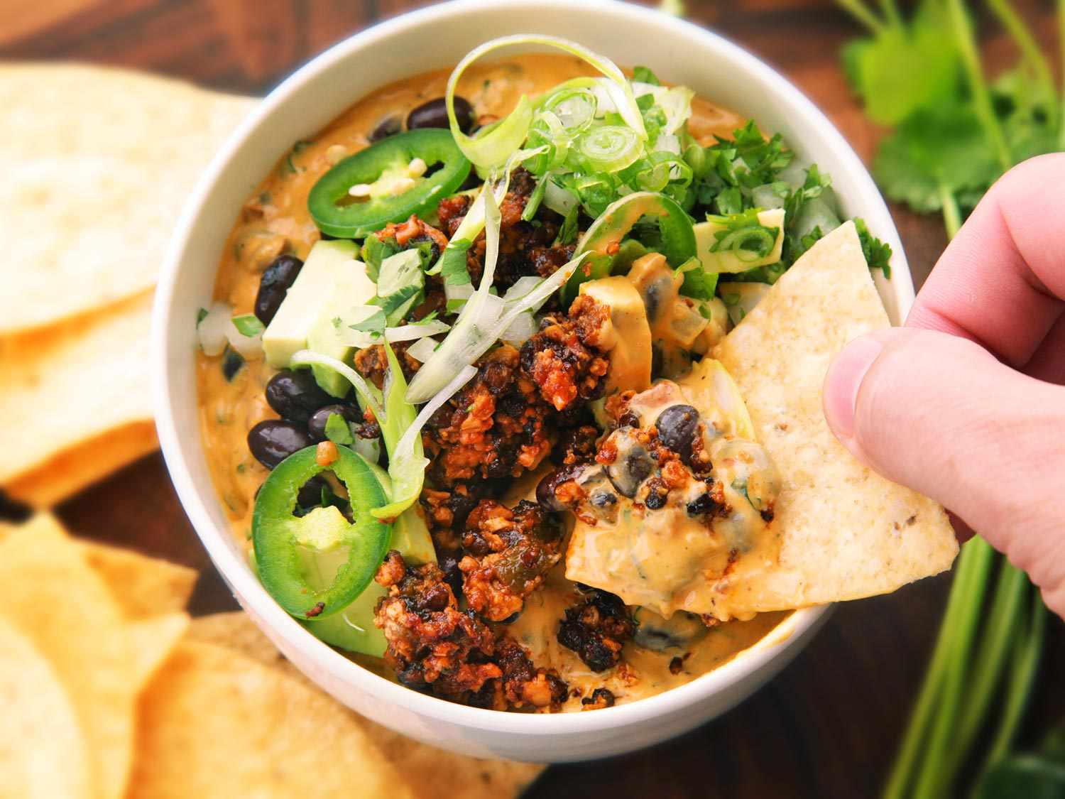 Handing hold a tortilla, digging into a bowl of fully-loaded vegan queso dip
