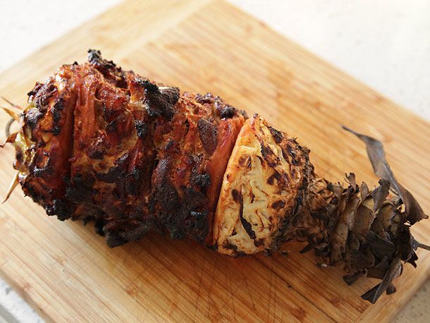 Grilled pork skewered inside a pineapple top and bottom sitting on a wooden cutting board.