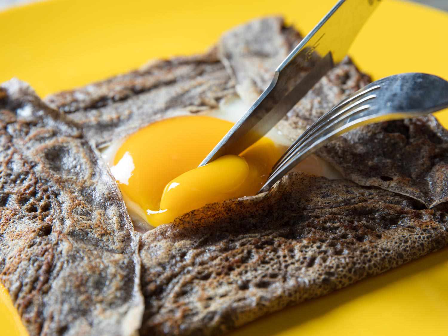 Close-up of a filled buckwheat crepe. A knife and fork are prodding the yolk of the exposed sunny-side-up egg. The yolk is not completely set, but it's more fudgy than it is runny.