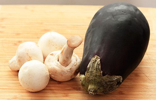Four whole white mushrooms sitting next to a whole eggplant on a cutting board.