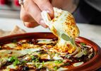A hand dipping a piece of bread into a baked cheese and herb dish served in a terracotta bowl