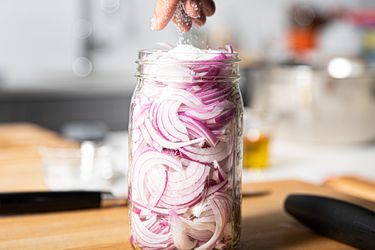 A hand adding liquid to a jar filled with sliced onions on a wooden countertop preparing quick pickled onions