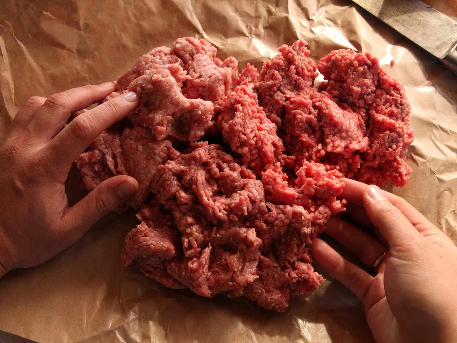 Ground beef being worked on by two hands on a brown paper surface.