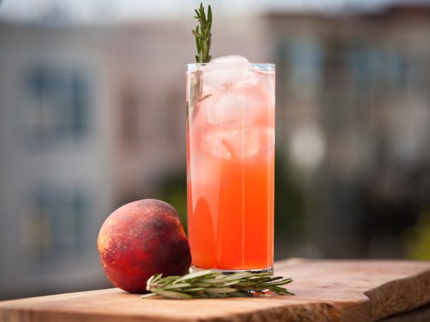 Profile view of a navy fizz cocktail, served in a tall glass with a rosemary sprig garnish. More rosemary and a ripe nectarine are nearby.