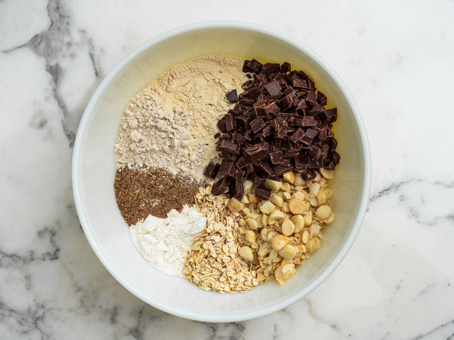 An assortment of dry ingredients for lactation cookies, arranged in a large bowl: chocolate chips, brewer's yeast, oat flour, all purpose flour, rolled oats, flax seed, macadamia nuts.