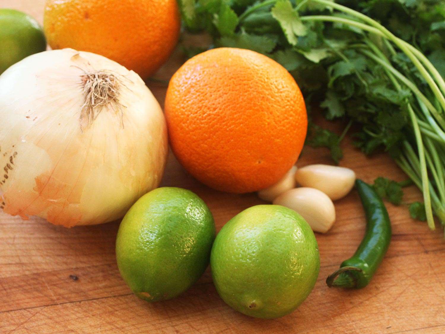 Onion, oranges, limes, garlic, serrano chile, and fresh cilantro on a wooden cutting board for a carne asada marinade.