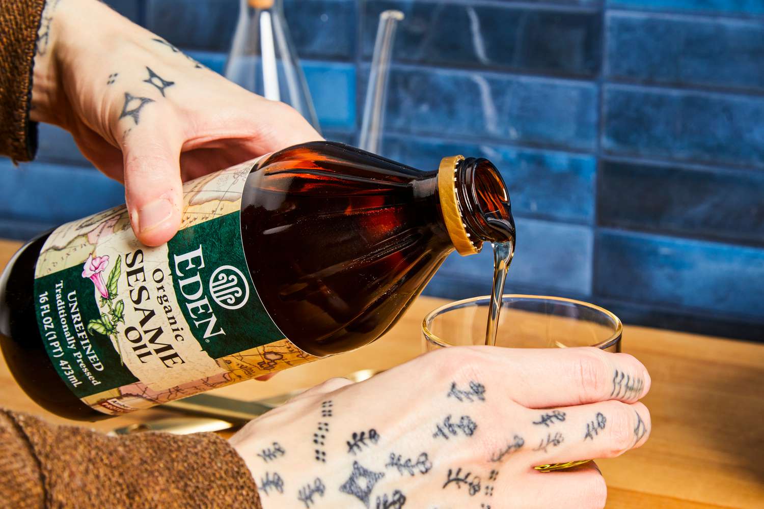 A hand pours sesame oil from a bottle into a glass container, blue tiled wall in the background