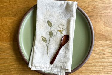 A ceramic plate with a cloth napkin and wooden spoon placed on top, featuring a leaf and flower pattern on the napkin