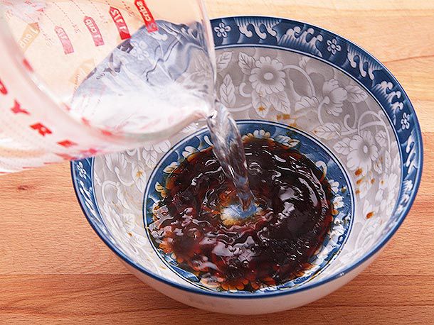 Some tare (soup base) is in a large floral blue bowl, and someone is pouring water from a glass measuring cup into the bowl. 