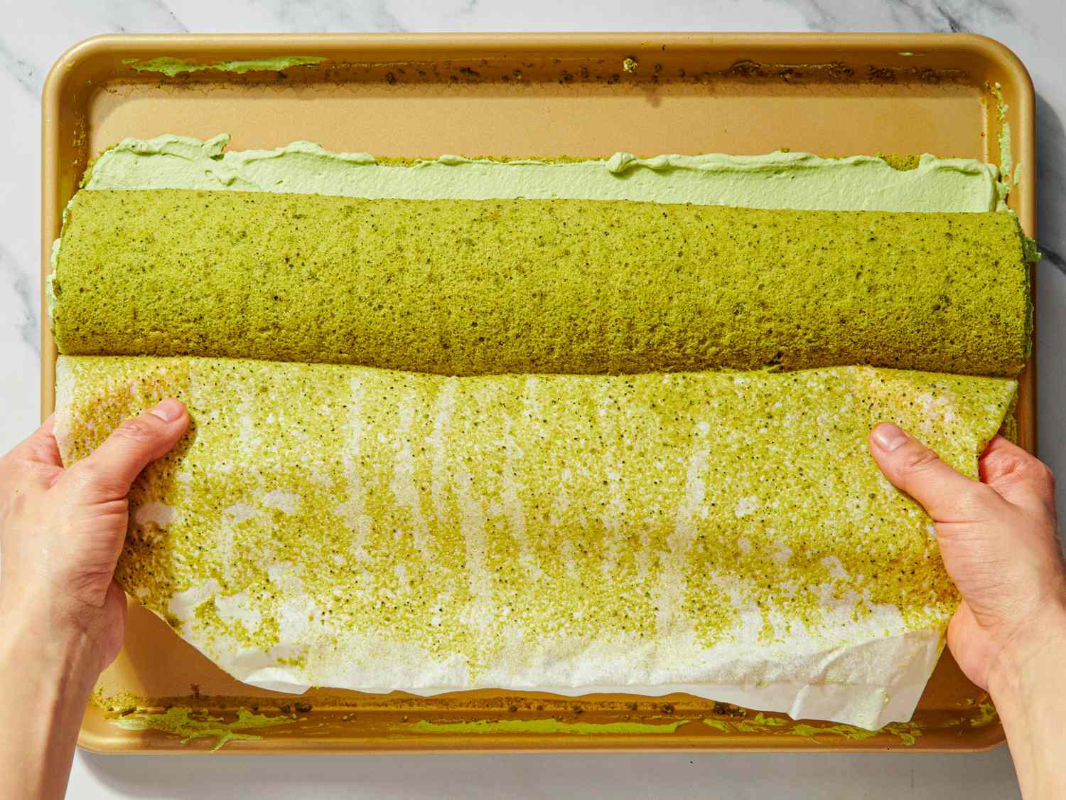 Hands rolling up a matcha swiss roll with icing on a baking sheet