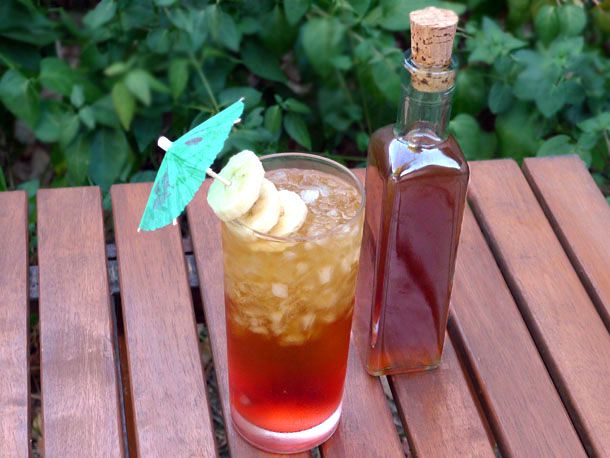 A bottle of DIY banana liqueur stands next to a tall cocktail garnished with an umbrella and banana slices.