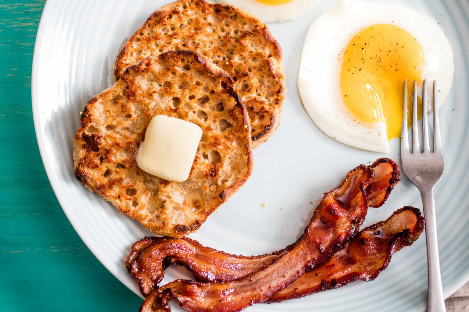 Toasted English muffin with butter, fried sunny side up eggs and bacon strips on a plate.