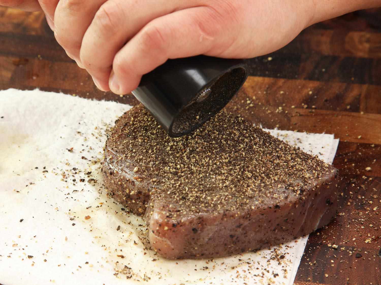 A cooked tuna steak has been laid on a paper towel, blotted, and is now getting another application of pepper.