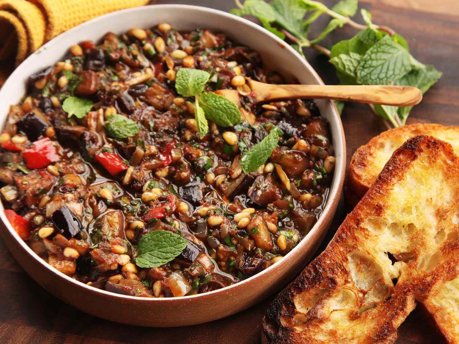 A bowl of Sicilian Eggplant and Pine Nut Caponata with fresh mint leaves. Two slices of toasted Italian bread are next to the bowl of caponata. 