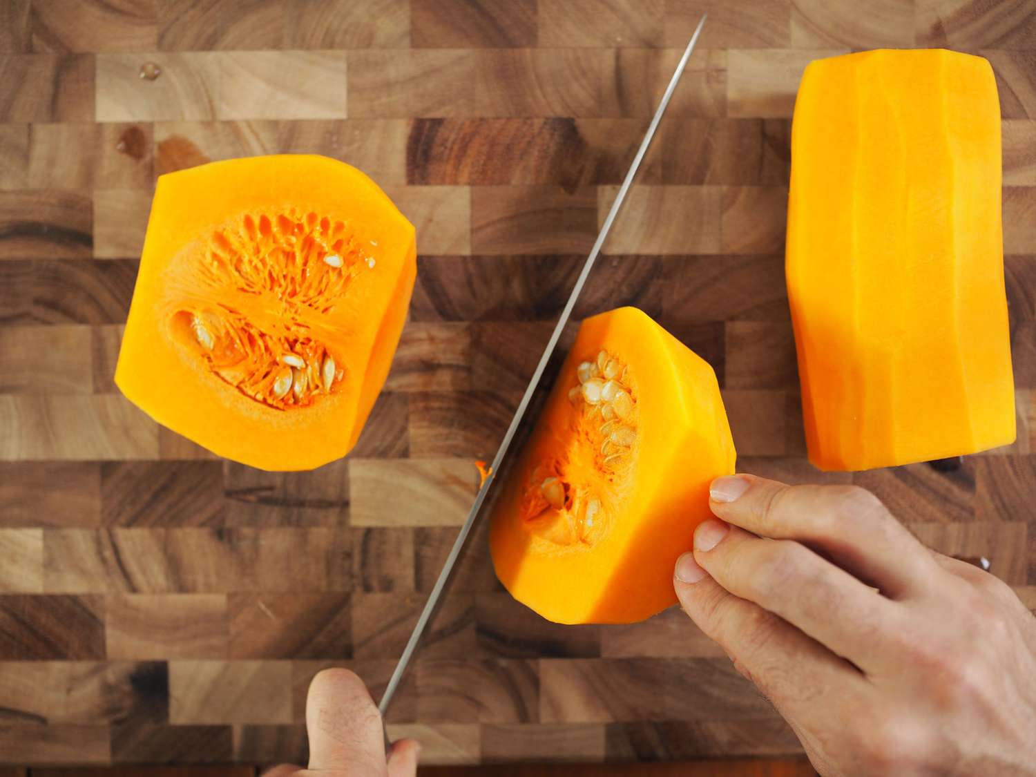 A butternut squash, being cut in half, on a cutting board. There are a pair of hands, one holding half of the squash and another holding the knife.