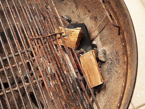 A small mound of half-lit charcoal placed along the wall of a kettle-style grill. Two pieces of hardwood have been laid on top and the grate has been fitted.