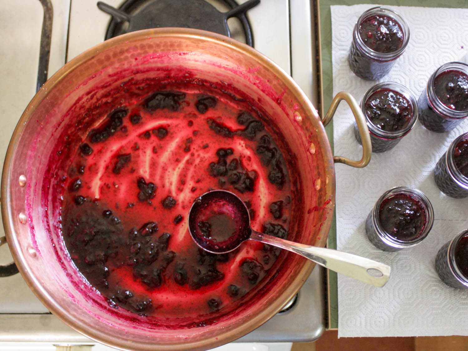 A copper put with a thin layer of cooked blueberry jam inside, with multiple glass jars filled with blueberry jam.