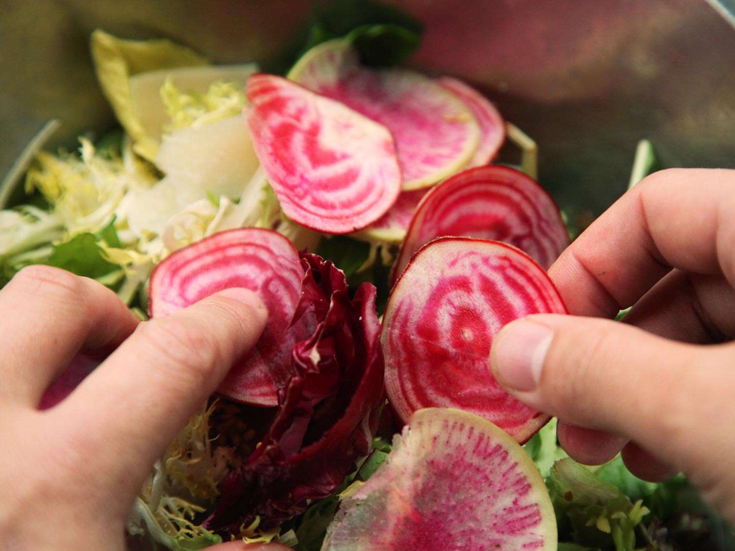 Shaved beets are added to a mixing bowl filled with a mixture of salad greens.