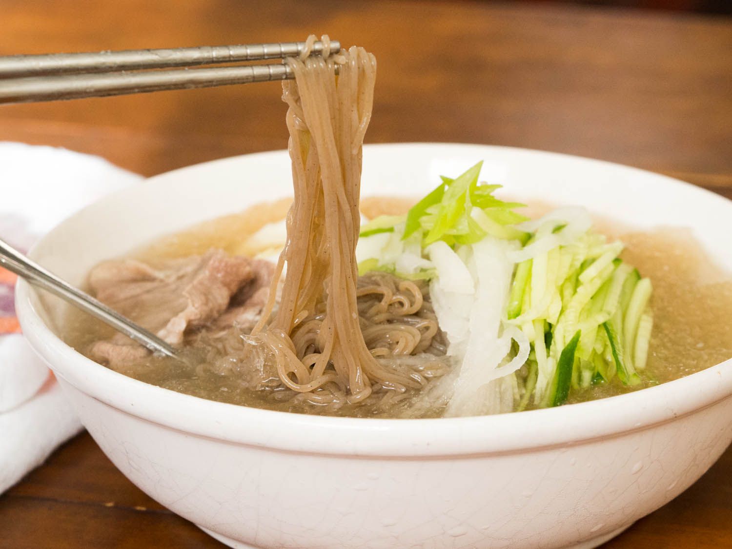 A pair of Korean metal chopsticks lifting cooked noodles out of a composed bowl of mul naengmyun