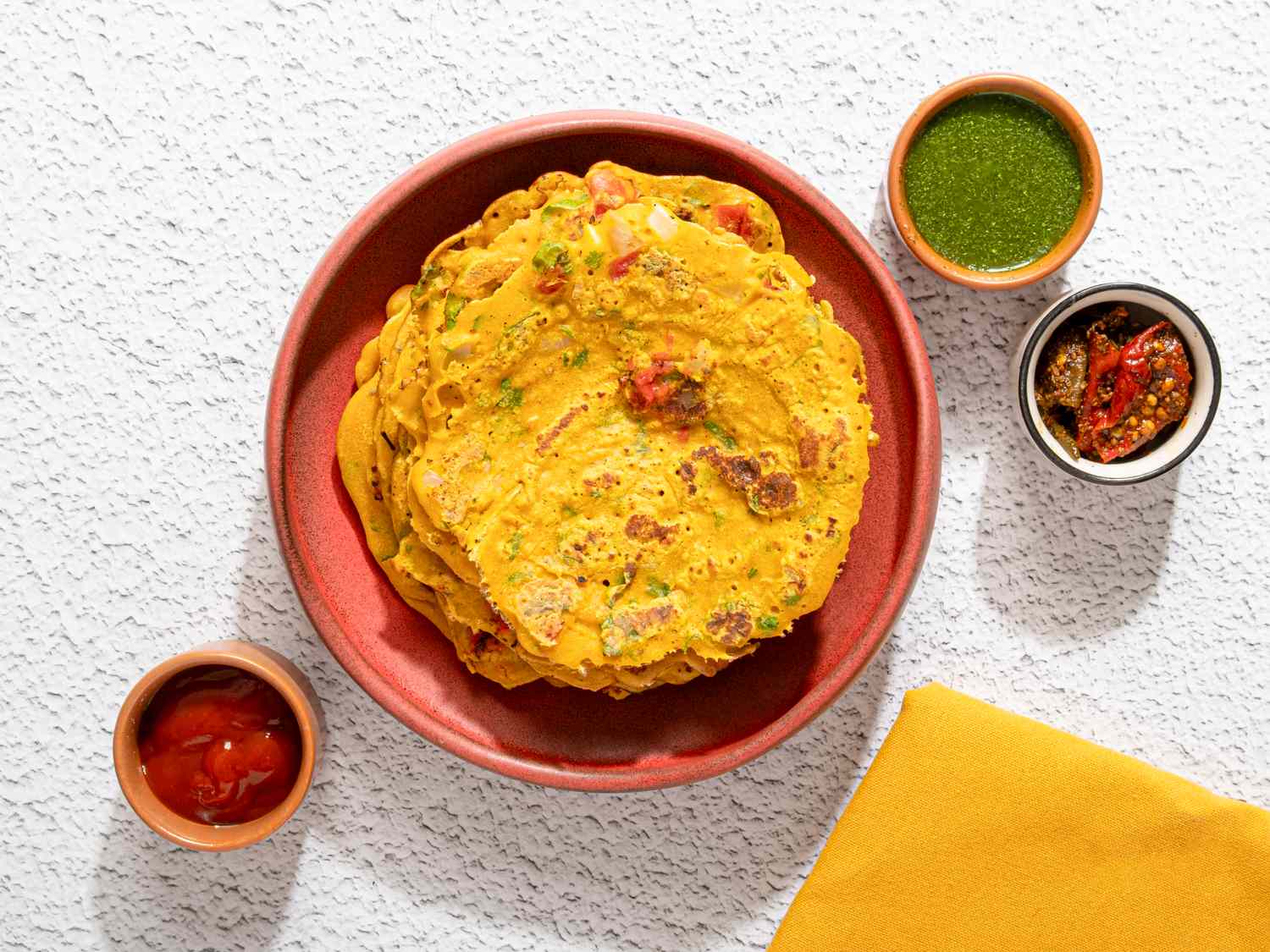 A dish of savory pancakes placed in a round plate accompanied by three small bowls of dipping sauces and a yellow napkin