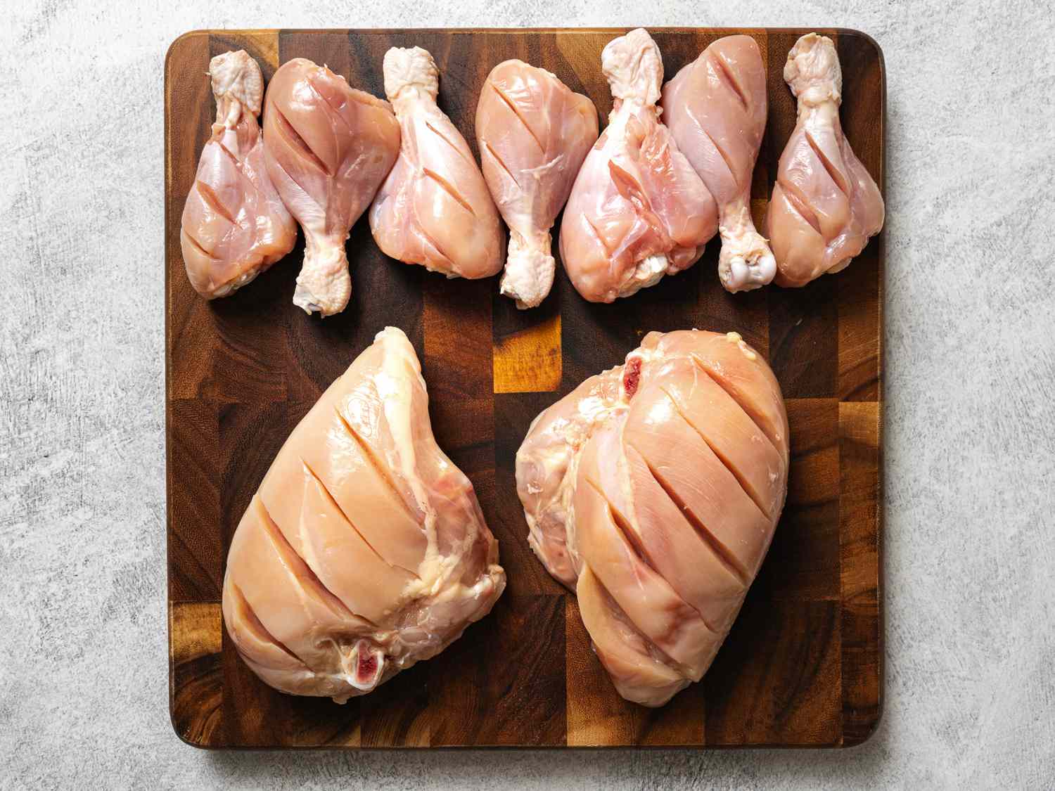 Two chicken breasts and seven chicken legs, the flesh scored with a knife, on a wooden cutting board.