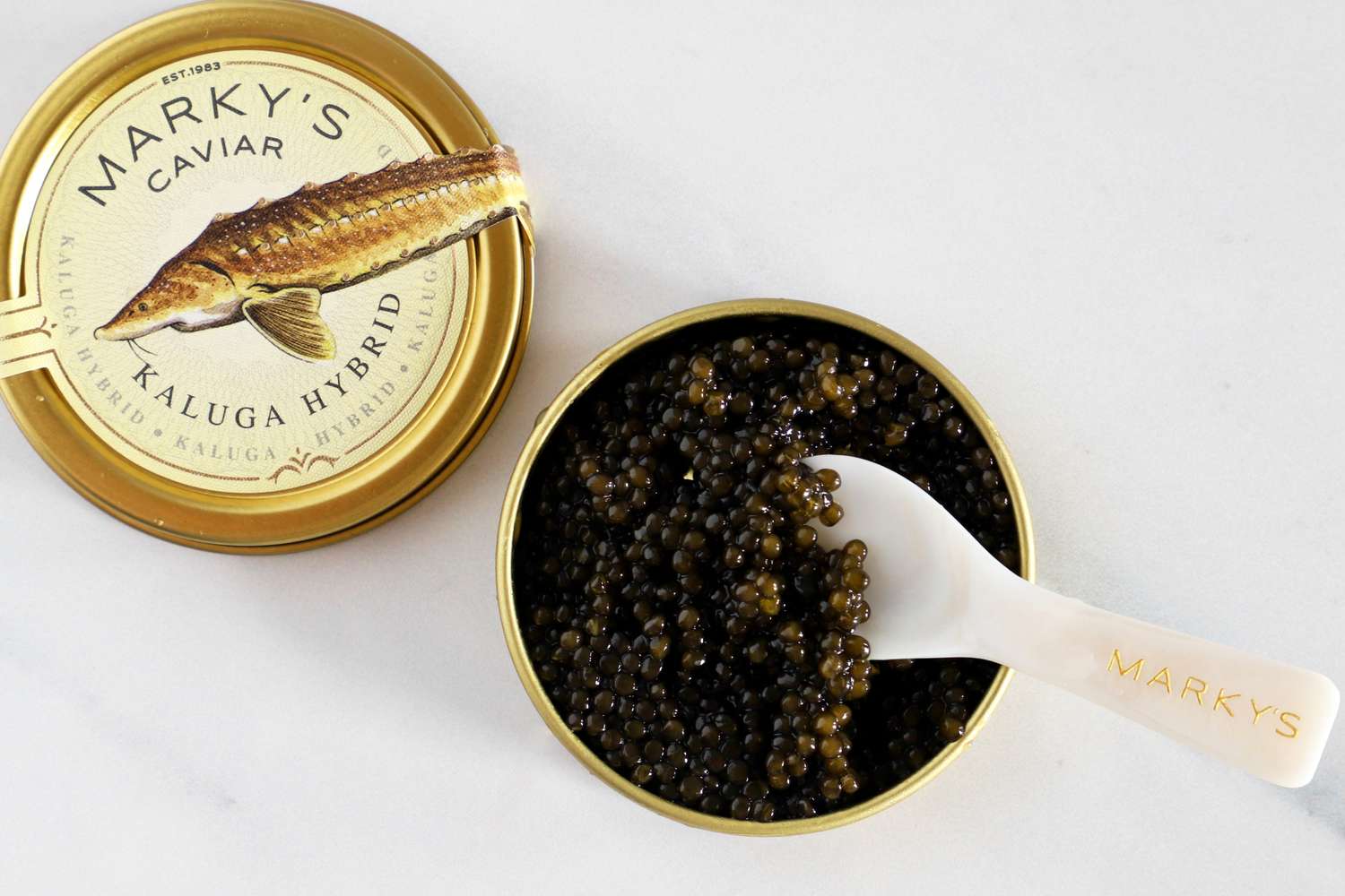 A tin of Markys Kaluga Hybrid caviar with a spoon containing caviar on a white surface