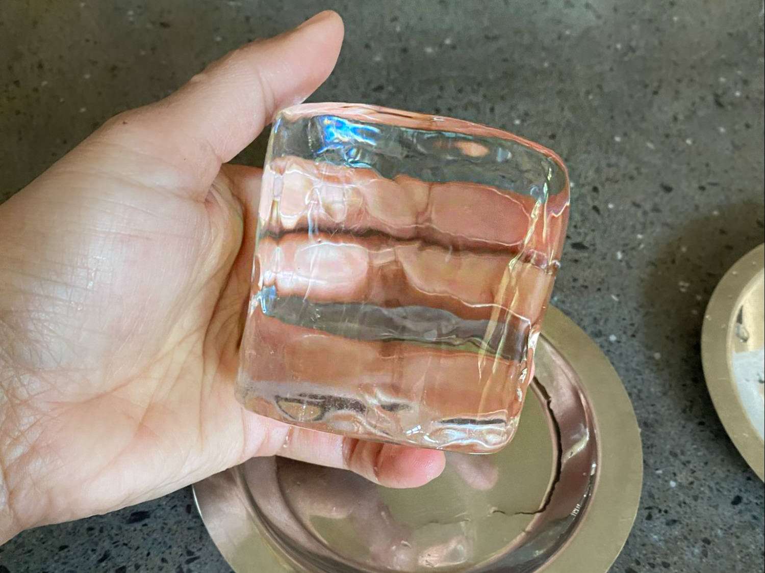 A hand holding a clear squareshaped ice cube over a reflective surface