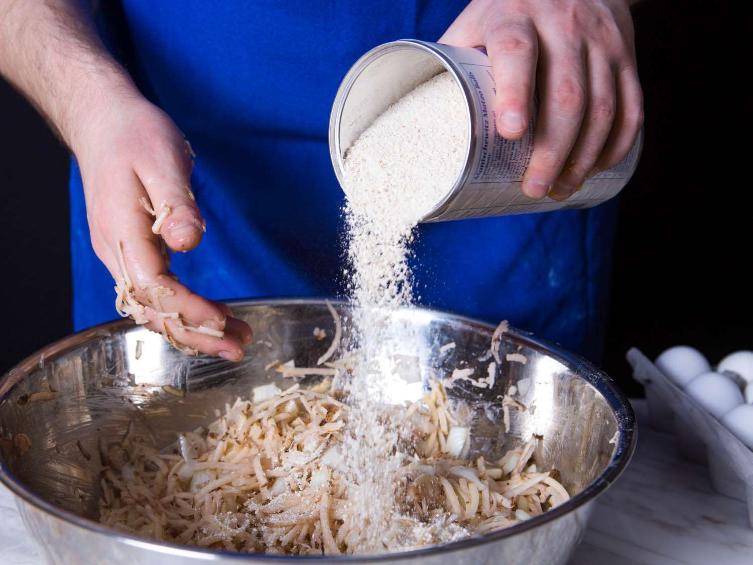 Matzo meal being poured into a large bowl with shredder potato mixture. 