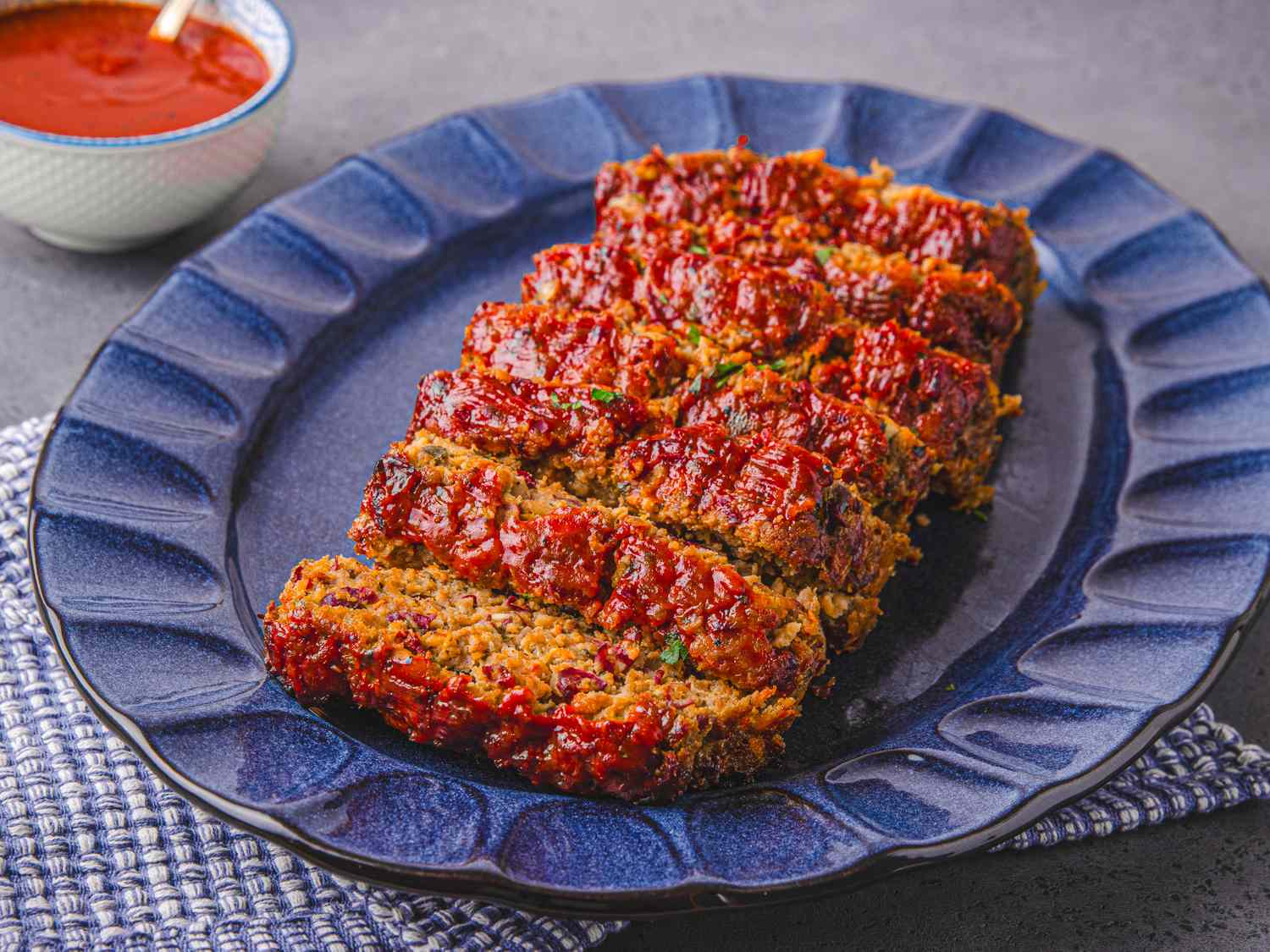 A dish with slices of meatloaf arranged on a blue serving plate a bowl of red sauce in the background
