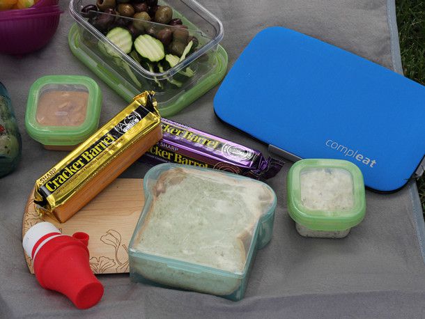 A small bamboo cutting board, two blocks of cheddar cheese, and containers of sandwich toppings on a gray picnic blanket. 