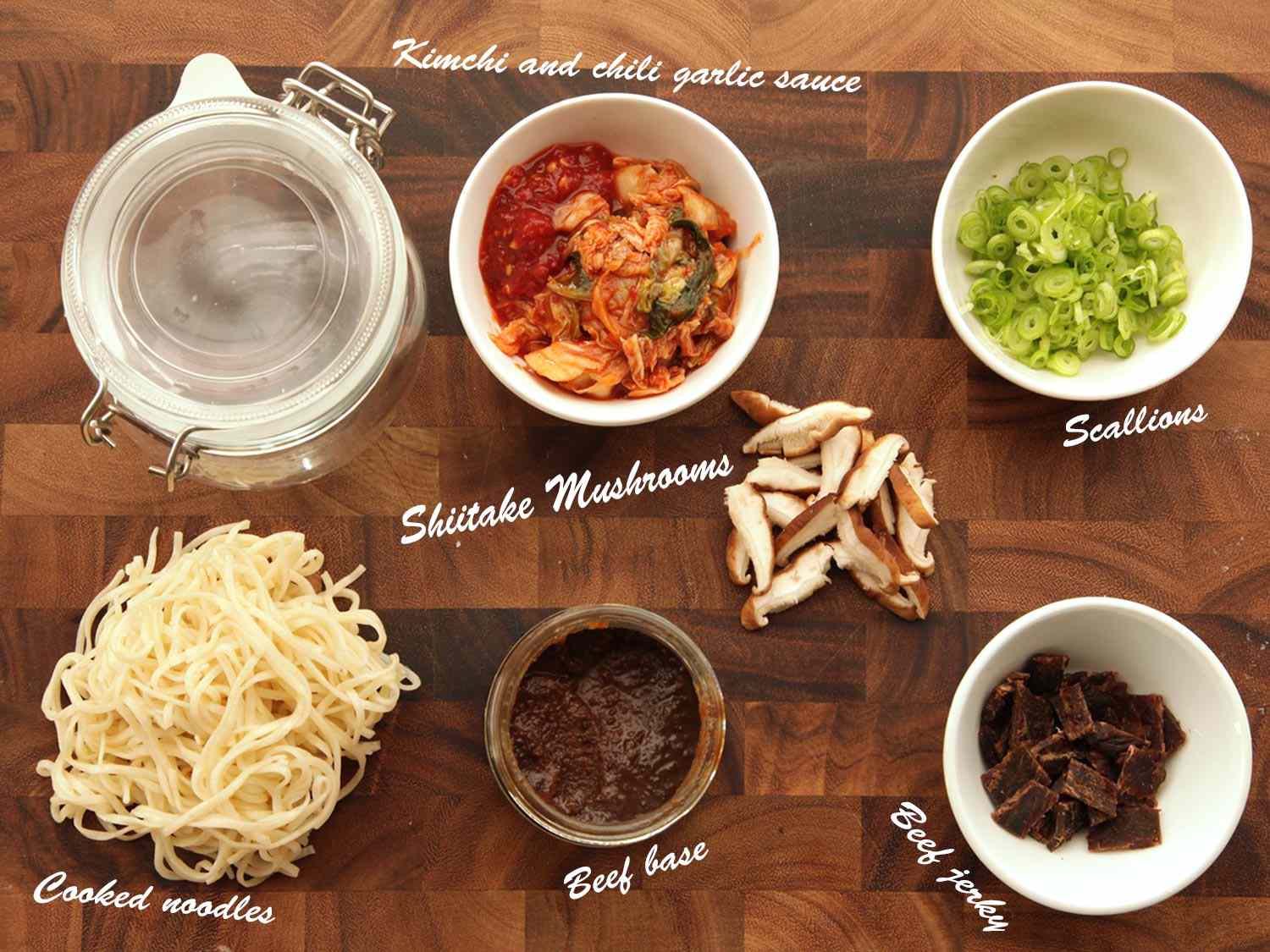 Overhead view of the ingredients needed to make spicy kimchi beef flavor instant noodles, arranged in bowls: kimchi and chile garlic sauce, shiitake mushrooms, scallions, beef jerky, beef base, and cooked noodles.
