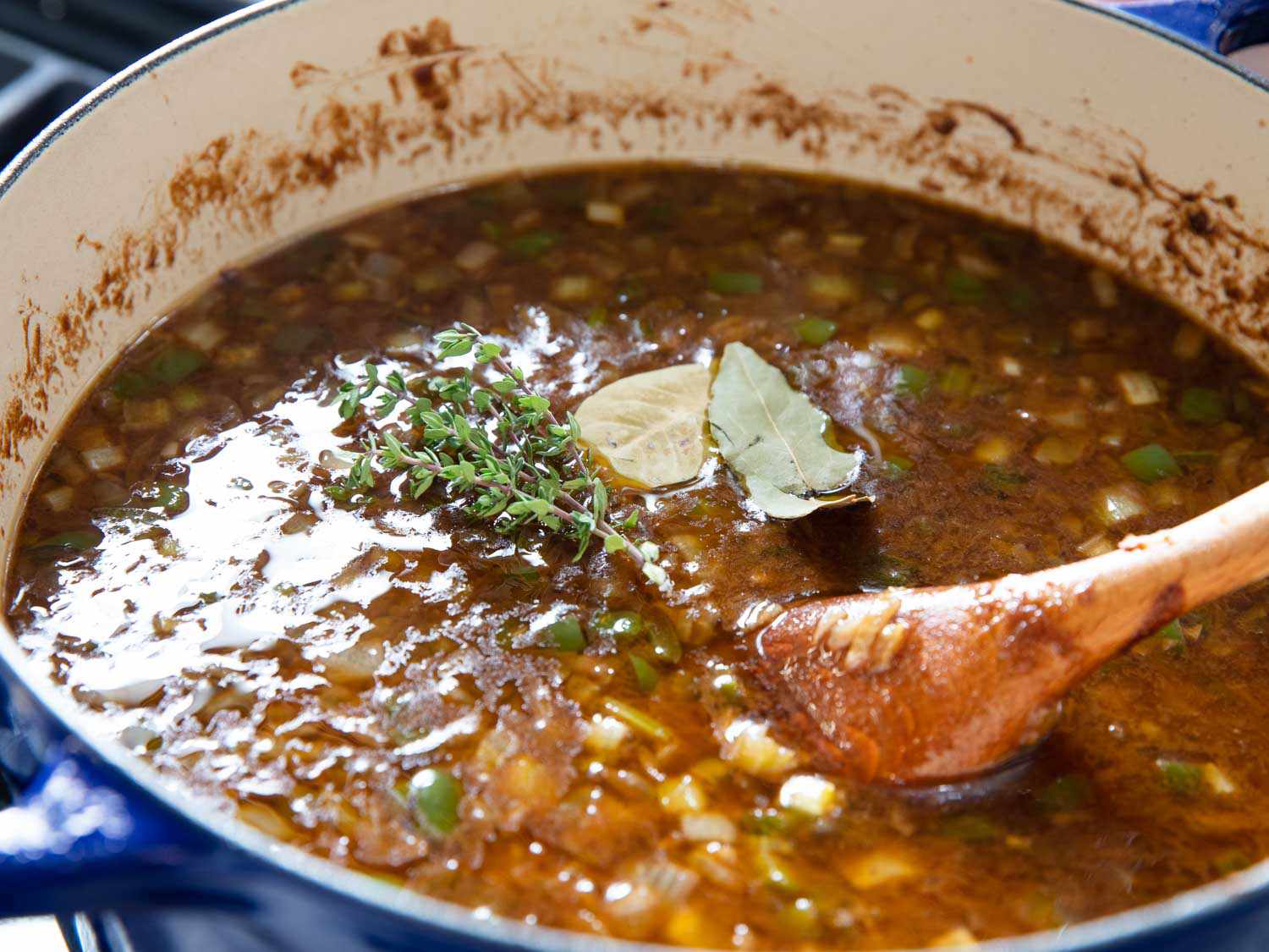 Stock, bay leaves, and thyme being added to the roux and stirred with a wooden spoon.