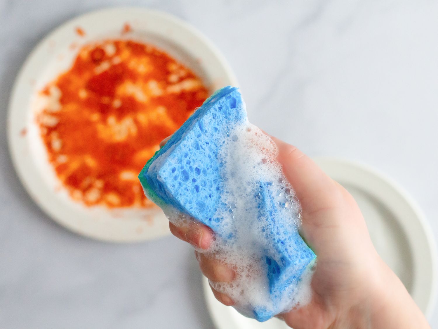 Hand holding a soapy sponge in front of a dirty plate