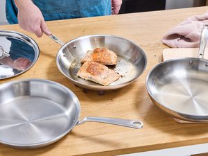 Three stainless steel skillets on a kitchen countertop. One has two cooked salmon fillets in it.