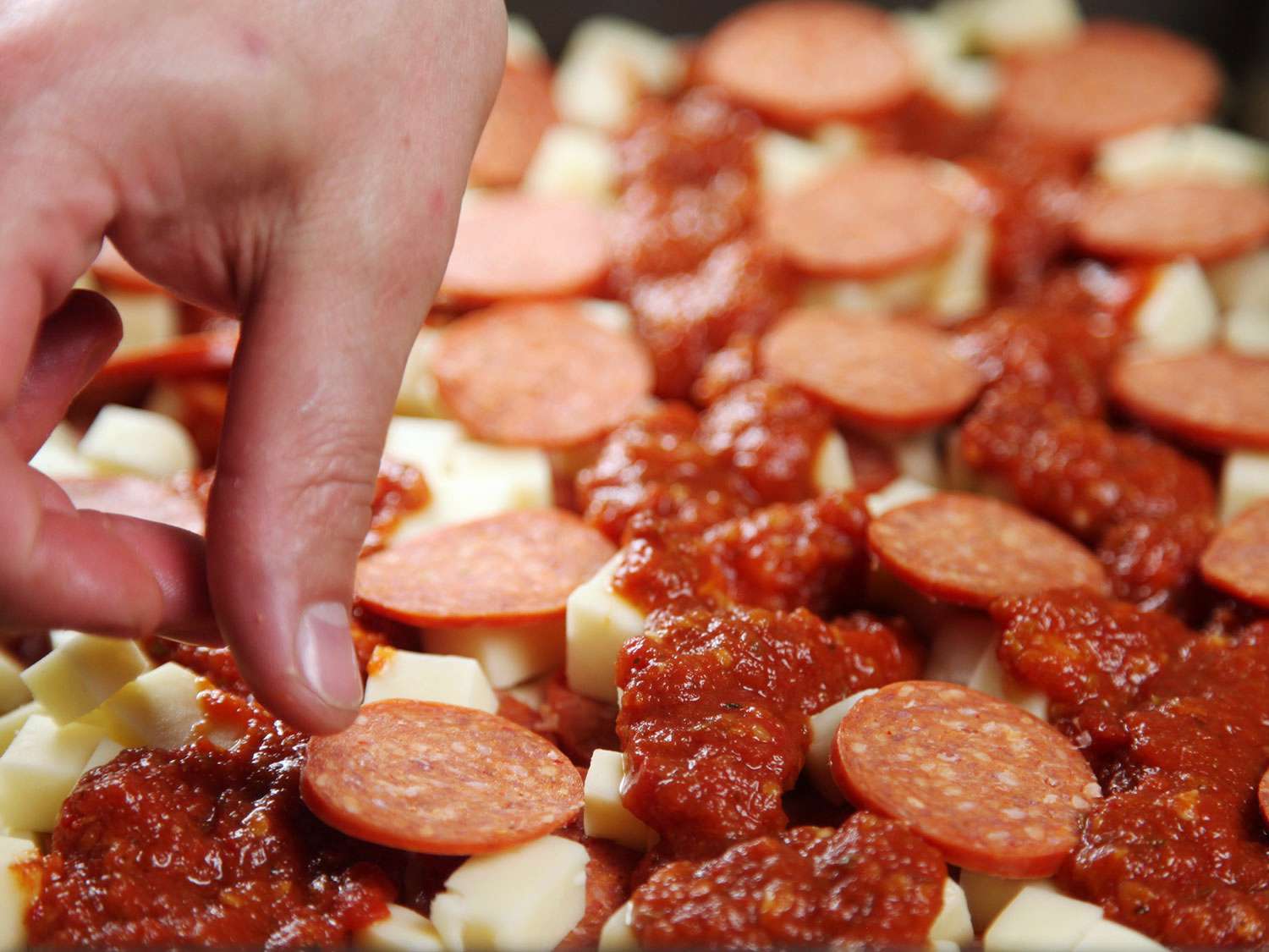 A hand placing pepperoni slices on top of the cheese layer of an unbaked Detroit-style pizza.