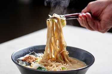 A hand lifting noodles with chopsticks from a bowl of ramen