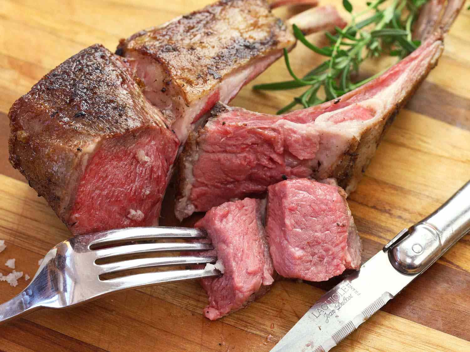 Sous vide rack of lamb on a wooden cutting board, with a fork stuck in it, next to a carving knife and rosemary sprigs