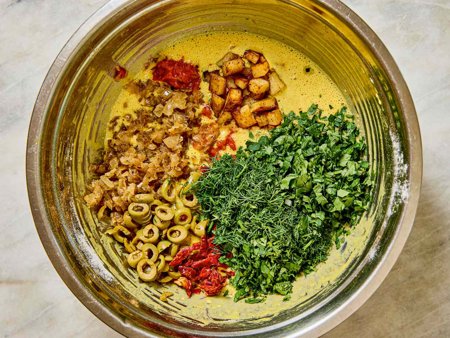 A large mixing bowl with ingredients like herbs olives and roasted vegetables divided into sections ready for mixing into a recipe